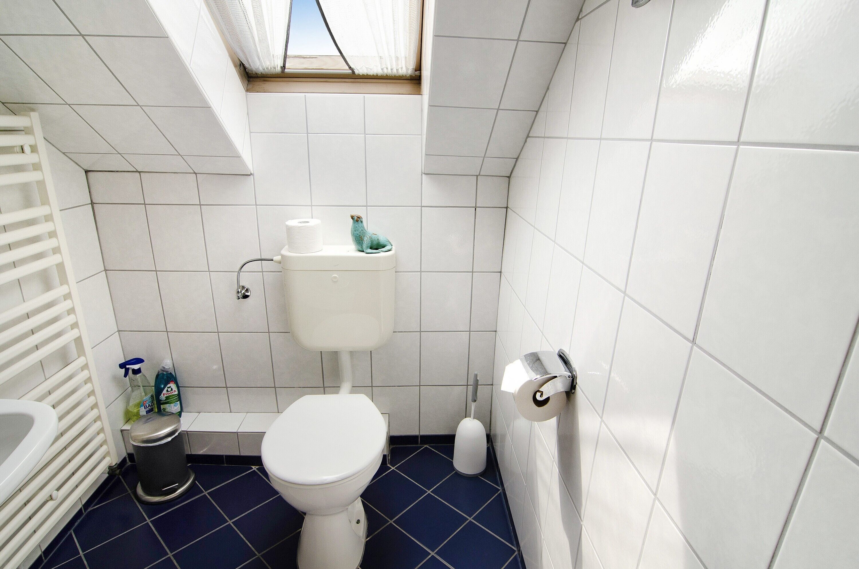 A bathroom with a toilet, a trash bin, a toilet paper holder, and a window.