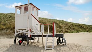 Ein Rettungsturm mit rot-weißer Gestaltung steht auf einem sandigen Strand, im Hintergrund Dünen.