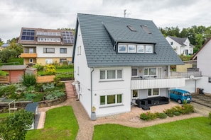 Une maison à deux étages avec un toit en pente, des panneaux solaires, un balcon et une voiture garée dans l’allée.