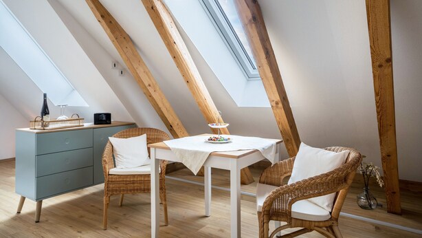 A room with a sloped ceiling, wooden beams, a dining table with chairs, a small cabinet, and a vase with flowers. - Apartment "Annele" with Private Terrace & Wi-Fi (Loßburg)