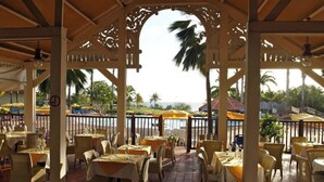 An outdoor dining area with tables and chairs, a view of the sea, and palm trees. - Studio privé dans le complexe de Pierre et Vacances (Sainte Luce)