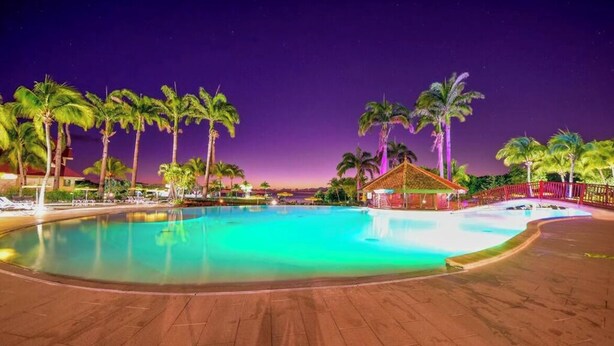 A nighttime swimming pool with palm trees and a small hut, surrounded by a wooden deck. - Studio privé dans le complexe de Pierre et Vacances (Sainte Luce)