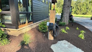 Un bâtiment moderne doté de grandes fenêtres, d’un poteau en bois muni d’un dispositif, et d’un espace jardin avec du paillis et des plantes.