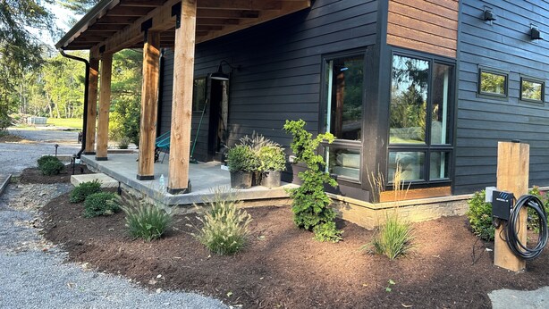 Une maison moderne dotée d’un porche abrité, d’un jardin avec différentes espèces de plantes et d’une borne de recharge pour véhicules électriques, bien visible.