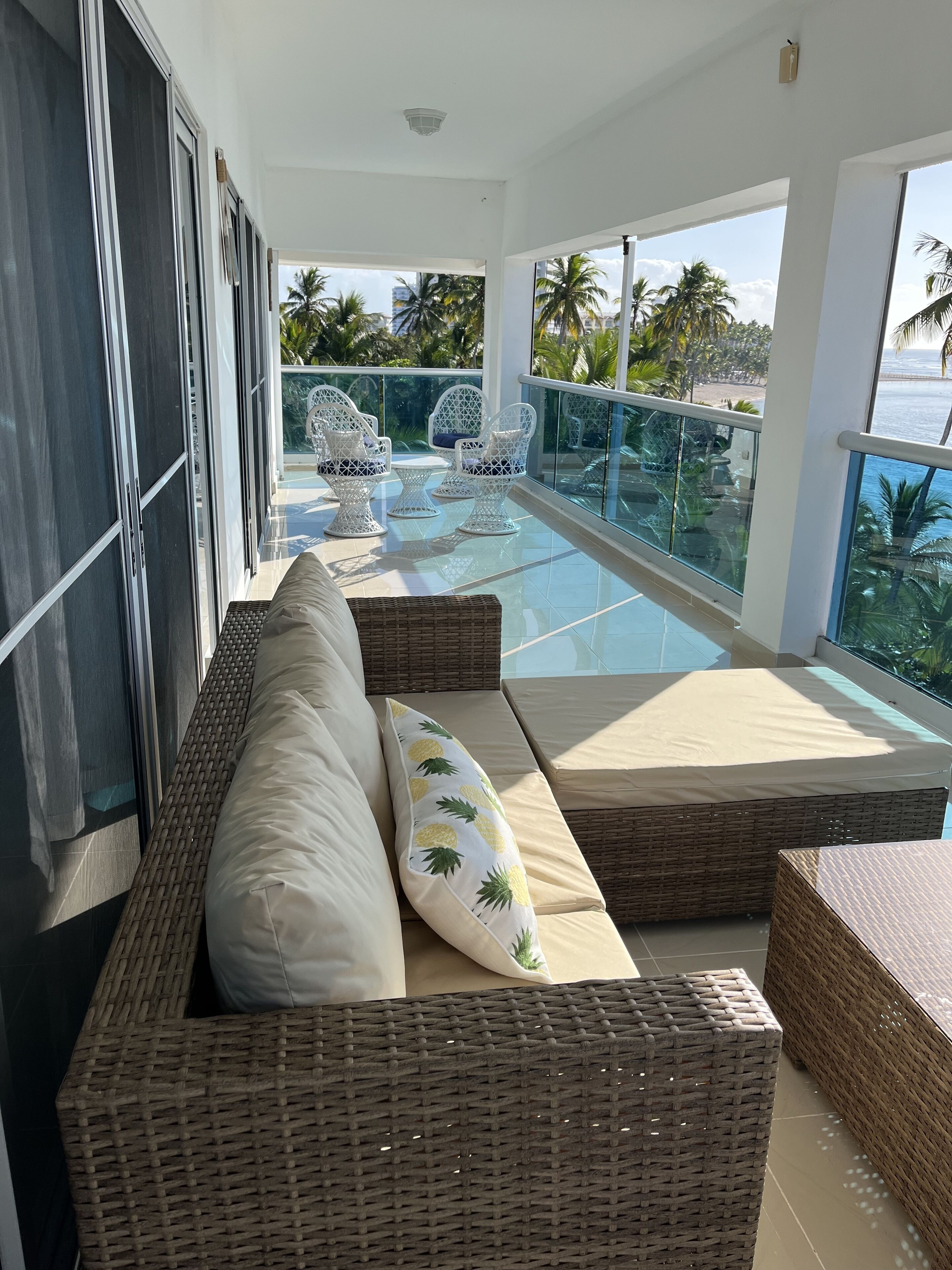 Een balkon met rieten meubels, een glazen hekwerk en uitzicht op palmbomen en de zee.