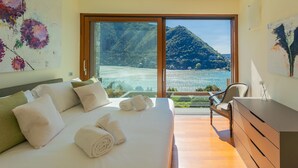 Ein Schlafzimmer mit einem großen Bett, einem Sessel, einem Schminktisch und Blick auf einen See und Berge.