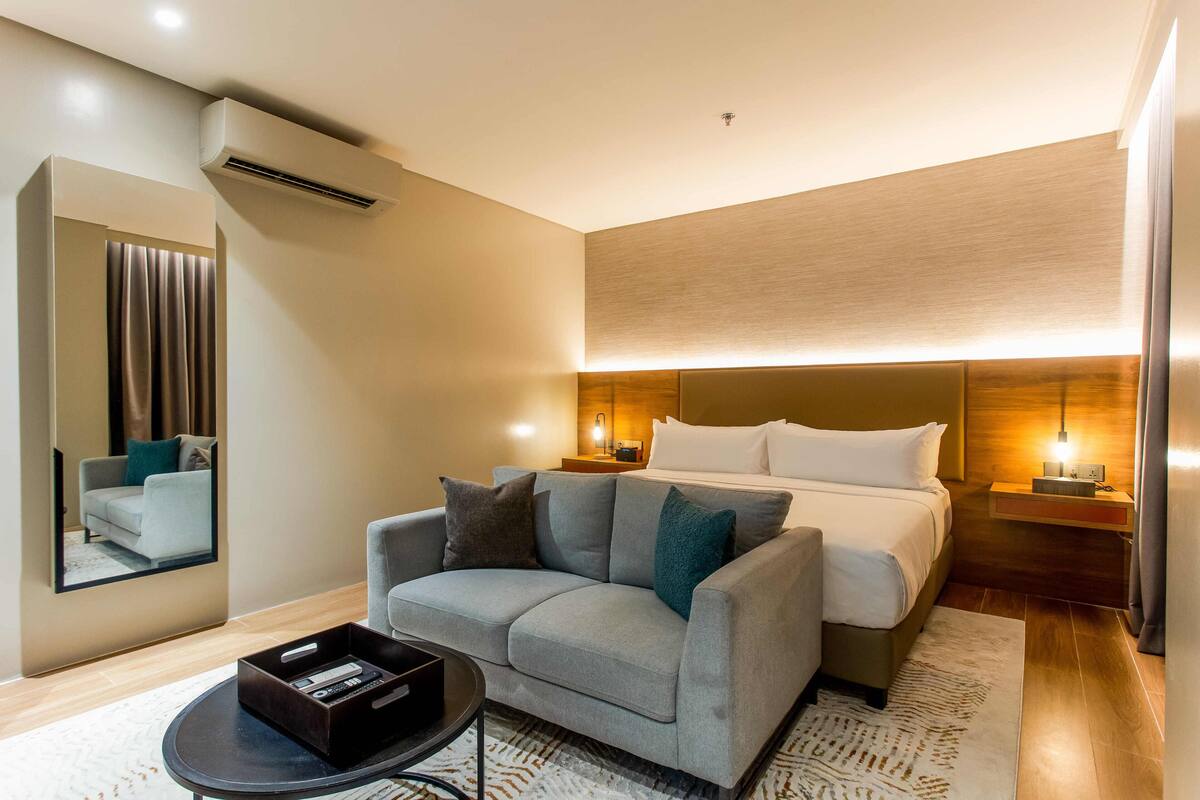 Ein modernes Hotelzimmer mit grauem Sofa, einem Bett mit weißer Bettwäsche, einem Nachttisch mit Lampe und einem großen Spiegel an der Wand.