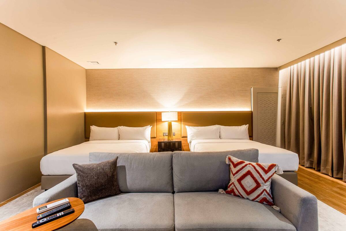 Ein Hotelzimmer mit zwei Betten, einem Sofa, einem kleinen Tisch und Fernbedienungen sowie einer Lampe.