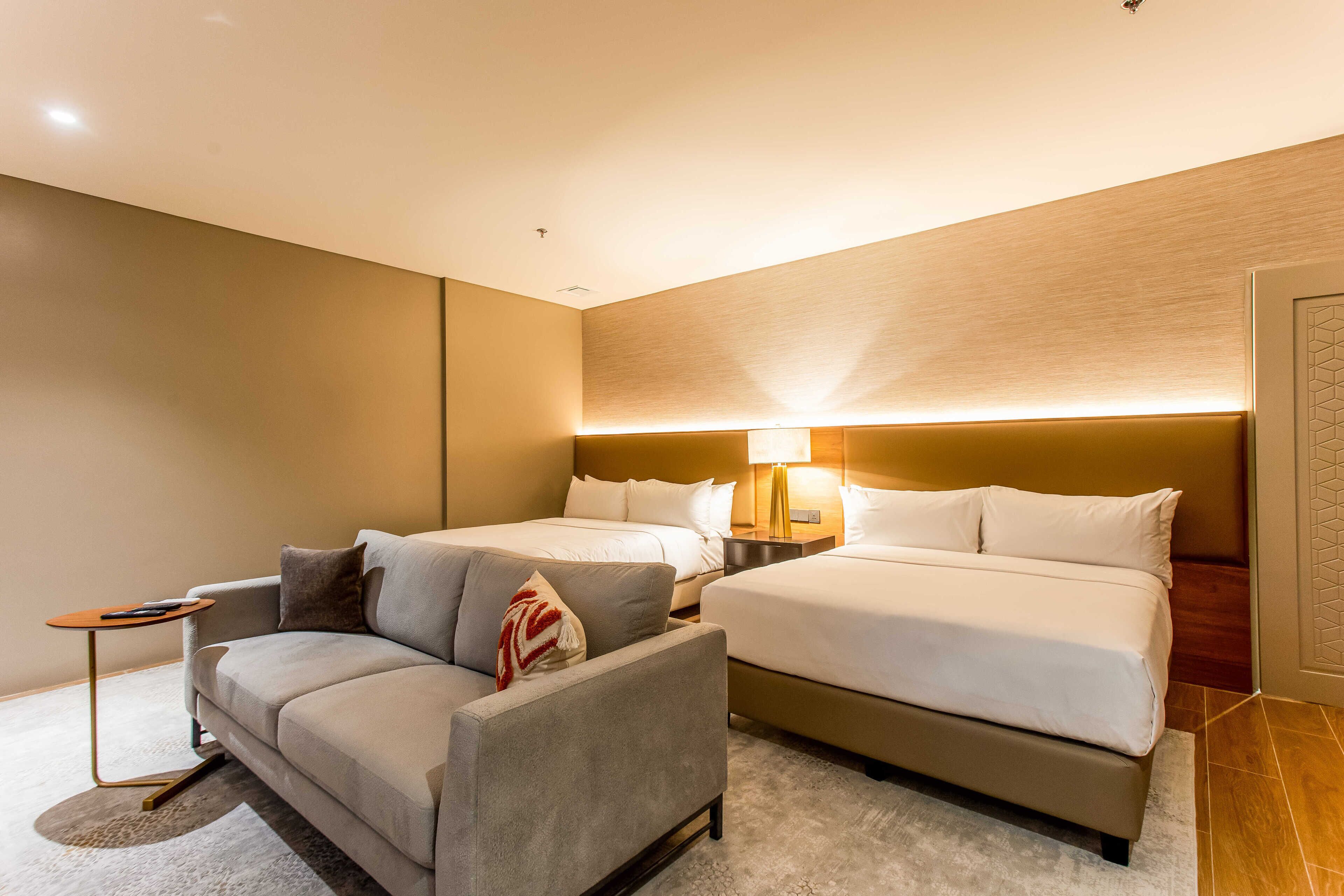 A hotel room with a sofa, two beds, a nightstand, and a lamp.
