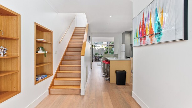 Un interior moderno con una escalera de madera, estanterías empotradas, una zona de cocina y un cuadro llamativo en la pared.