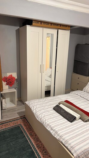 Une chambre à coucher avec un lit, une armoire, une petite table sur laquelle est posé un vase, et un tapis à motifs.