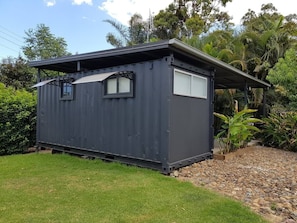Situated in a serene garden, this compact structure offers a peaceful retreat with large windows and a gravel path.