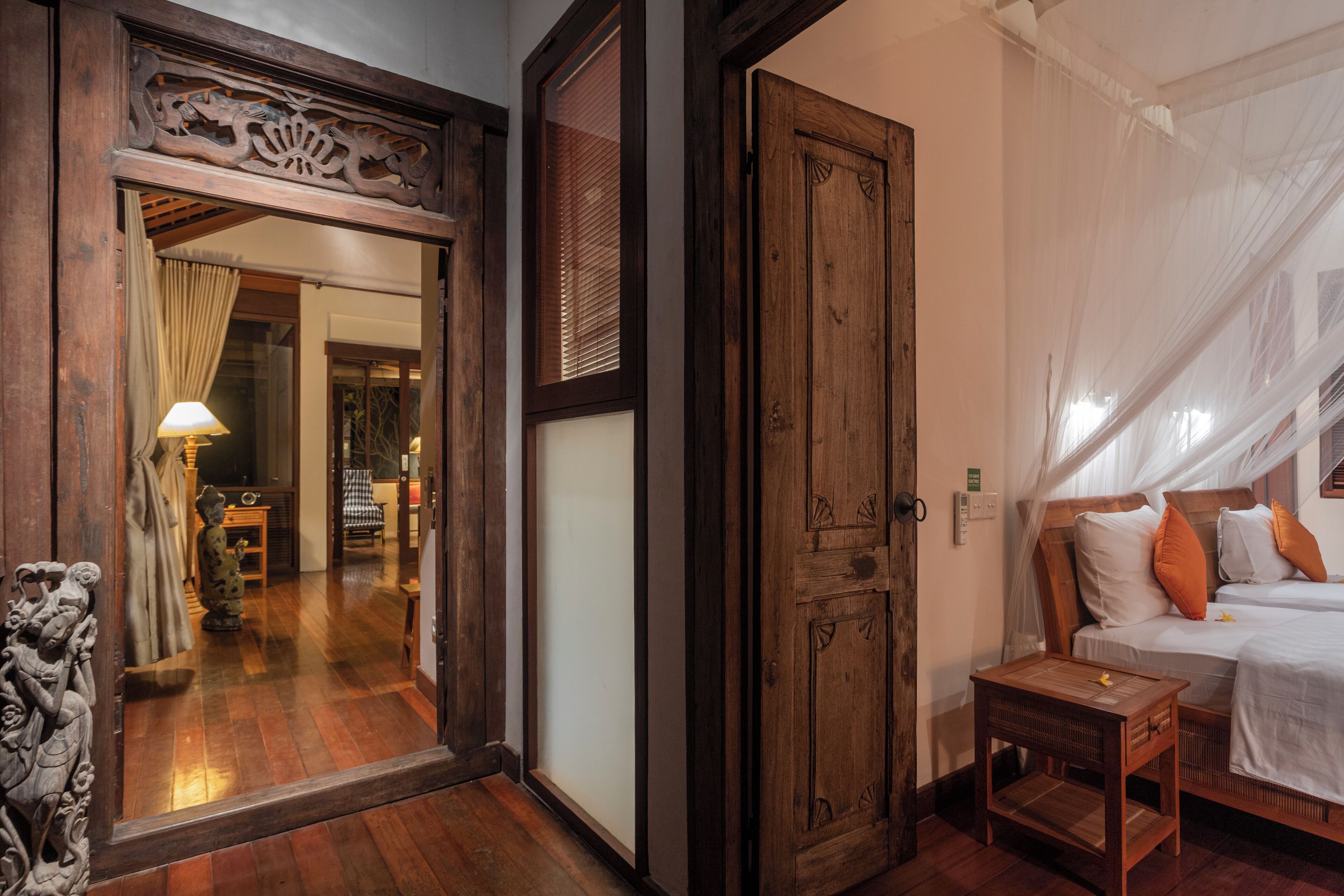 Una habitación tradicional con una puerta de madera, una cama con almohadas blancas y anaranjadas, y una mesita de noche de madera.