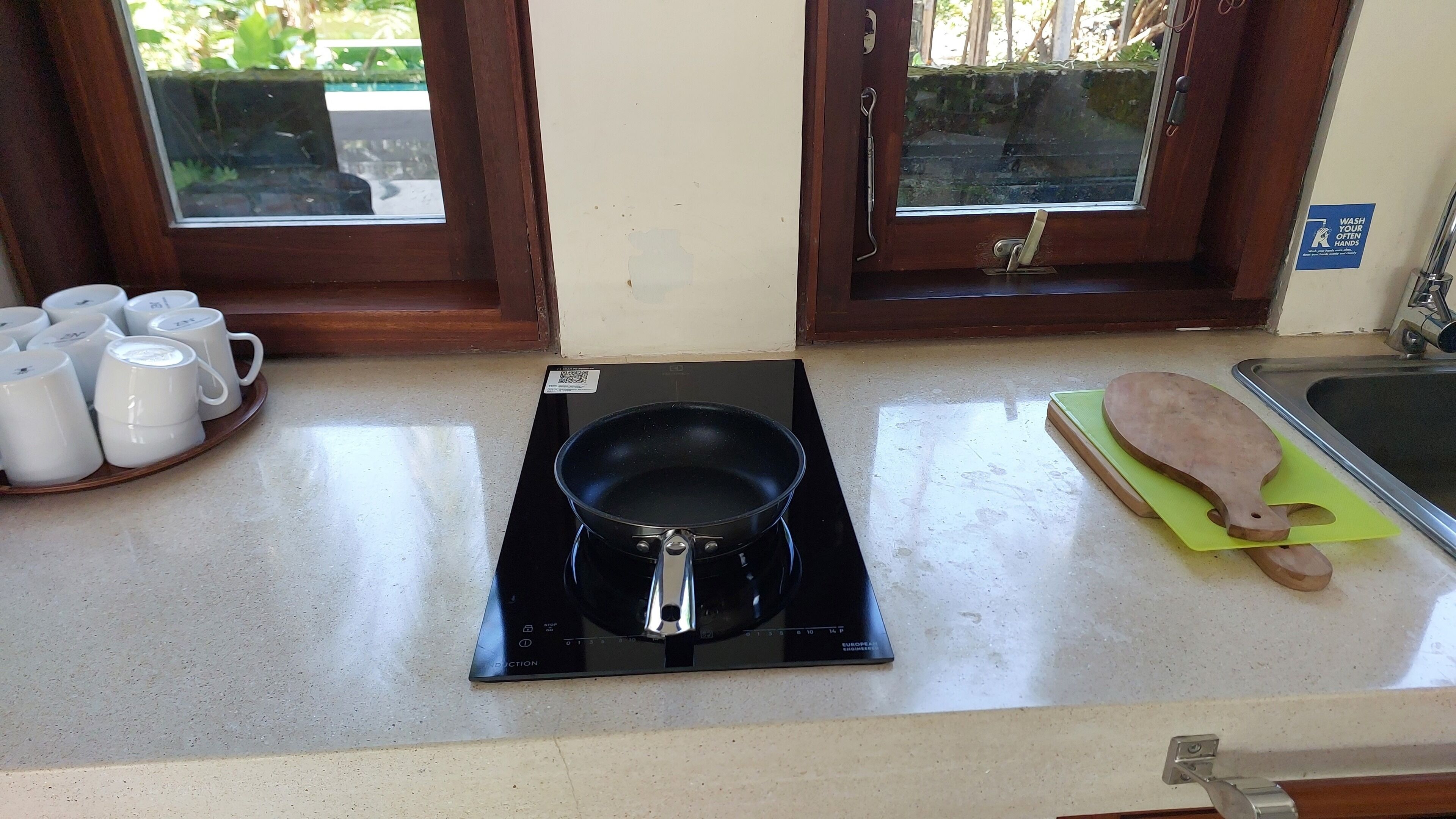 A kitchen area with a black induction cooktop, a tray of white mugs, a wooden cutting board, and a sink.