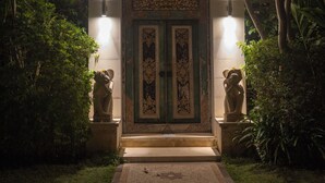 Una entrada nocturna con un camino que lleva a una puerta, flanqueada por dos jarrones grandes y rodeada de abundante follaje.