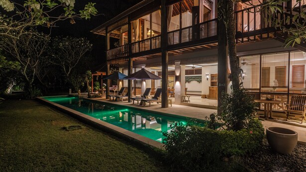 Un edificio de dos pisos con piscina, mobiliario exterior e interiores bien iluminados.