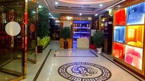 A hotel lobby with a circular emblem on the floor, glass doors, potted plants, and a brightly lit reception area.