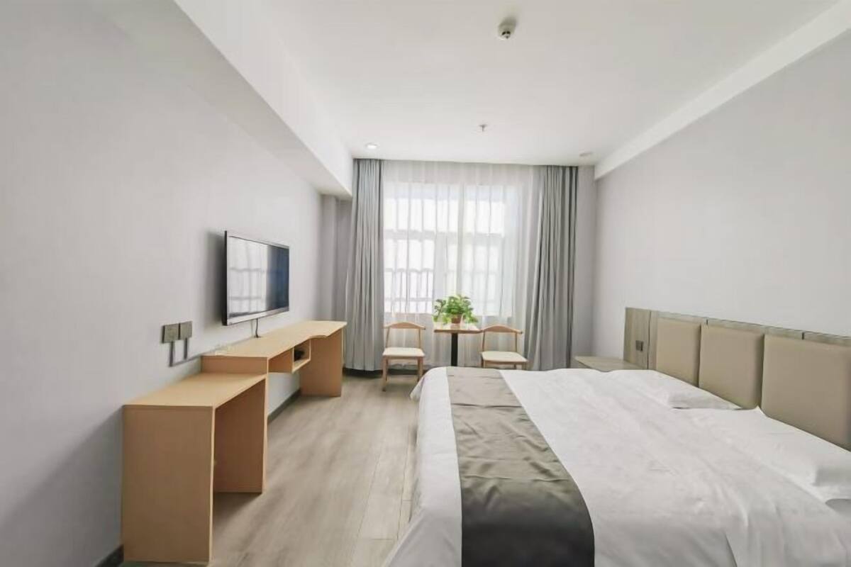 A hotel room with a bed, a desk, two chairs, a television, and a window with curtains.