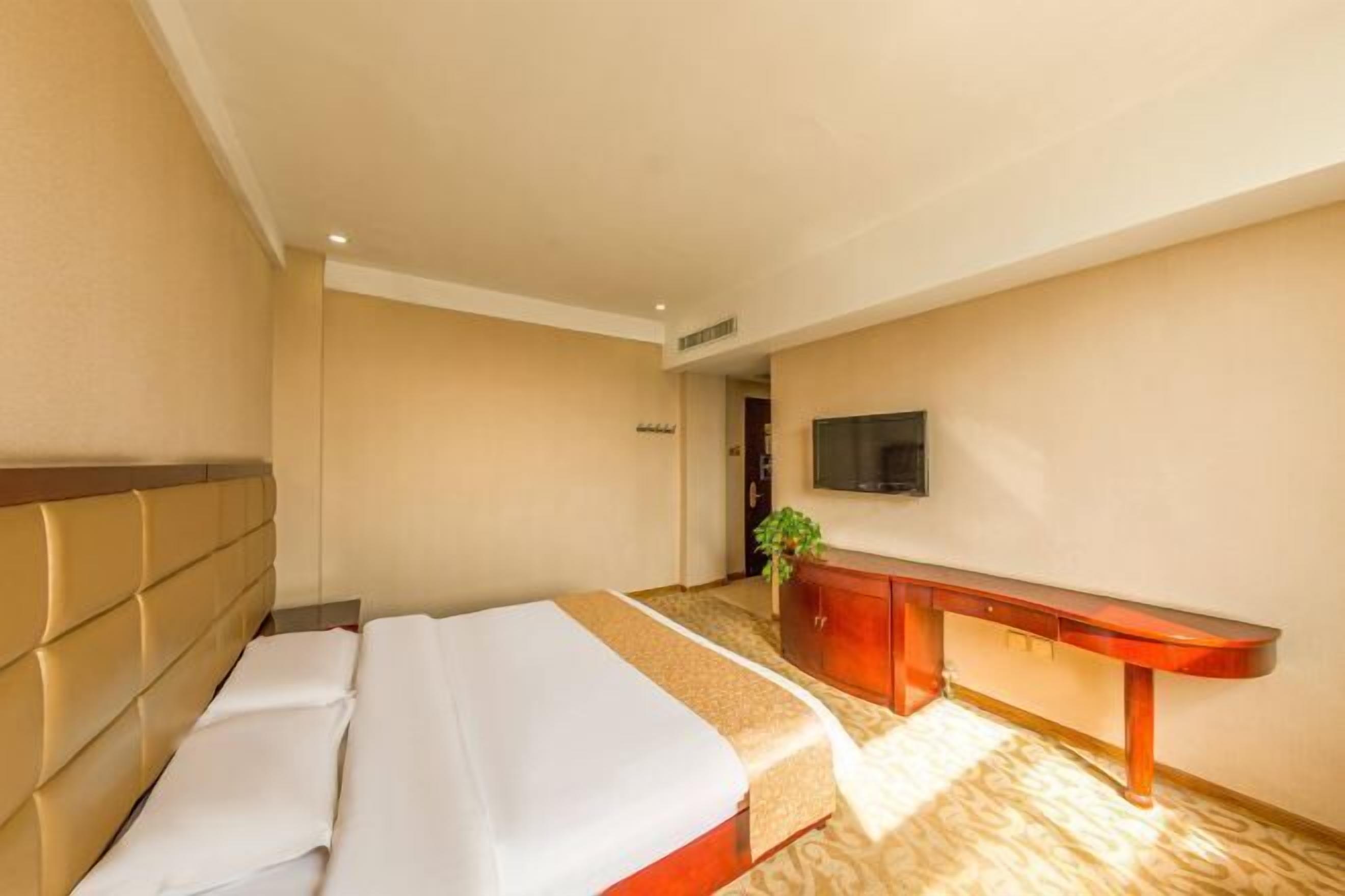 A hotel room with a bed, a wooden bench, a television, and a plant.