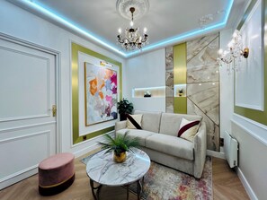 Un salon moderne avec un canapé, une table basse ronde, un pouf rose, un lustre et un grand tableau abstrait.