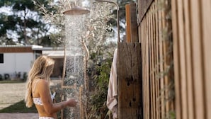 Une personne utilise une pomme de douche dans un espace extérieur, avec une clôture en bois et de la verdure en arrière-plan.
