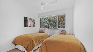 Ein Schlafzimmer mit zwei Betten, einem Deckenventilator, einem Fenster mit Blick auf Bäume und einem Bild an der Wand.