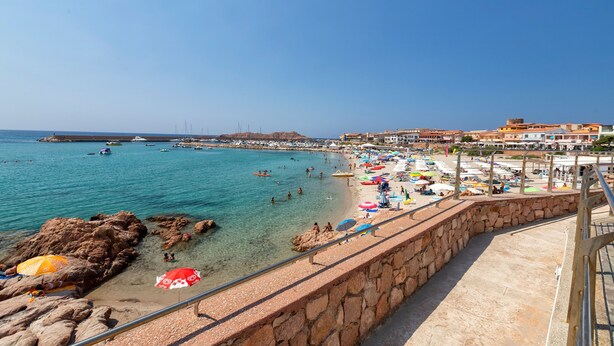 A coastal beach scene with clear water, rocky areas, and a stone wall.
