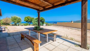 An outdoor patio with a wooden table and benches, a clear view of the sea, and a covered area with a wooden roof.