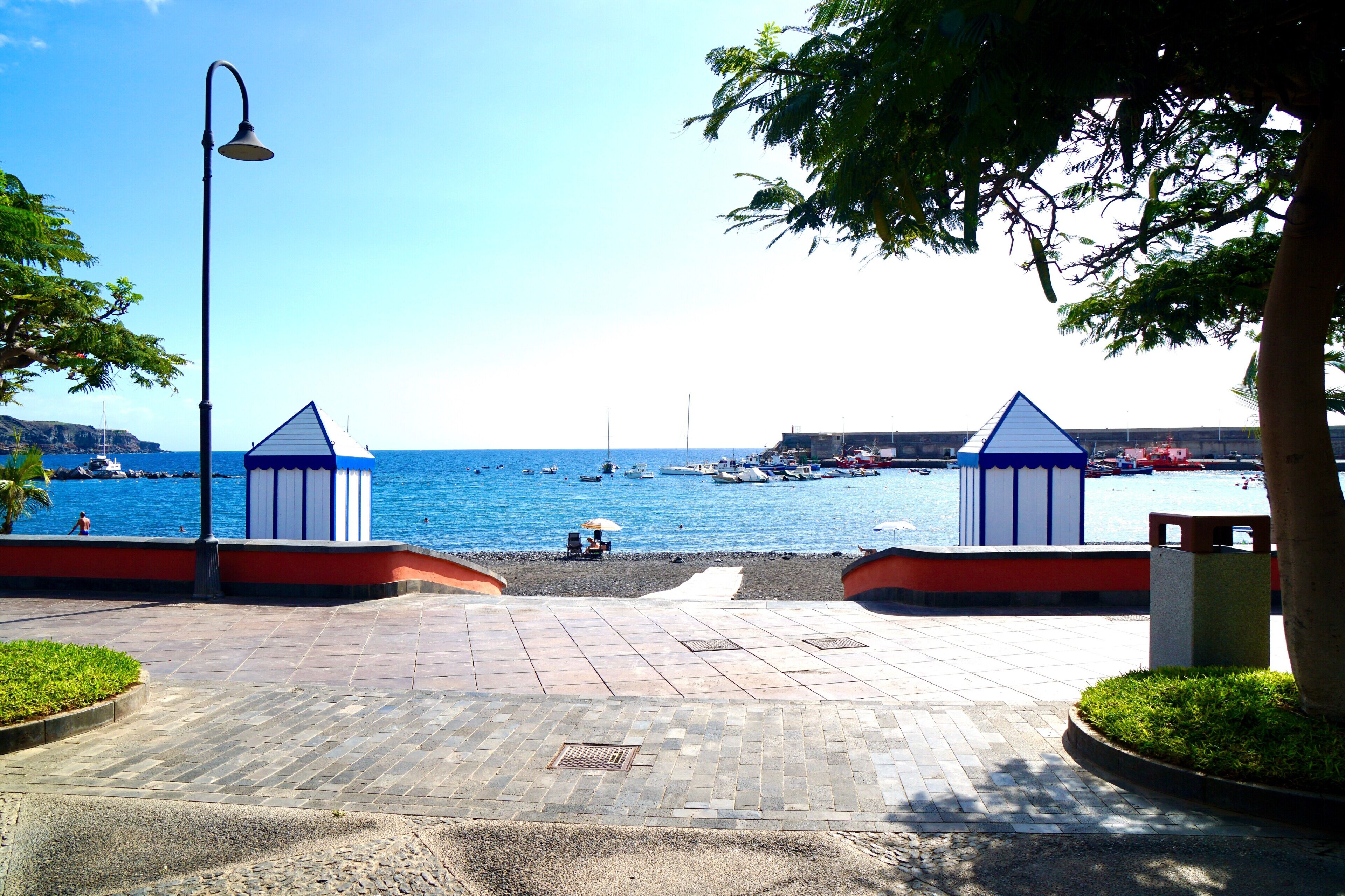 A paved walkway leading to a beach with two blue and white beach huts, a lamp post, and a clear view of the sea.