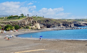 A beach with rocky terrain, a clear blue sea, and a distant view of a hill with buildings. - Holiday Apartment "Casa Emma" with Balcony & Wi-Fi (Playa de San Juan)