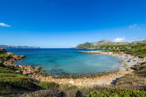 A coastal beach with clear water, rocky shore, and distant hills.
