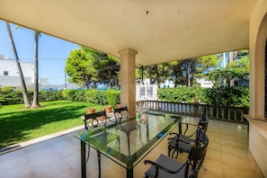 Un espacio comedor al aire libre con techo, que incluye una mesa y sillas de vidrio, rodeado por un jardín y vistas a edificios.