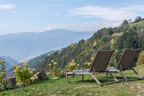 Deux fauteuils confortables sont placés sur une colline herbeuse, d’où l’on peut admirer un paysage de montagnes couvertes de forêts.