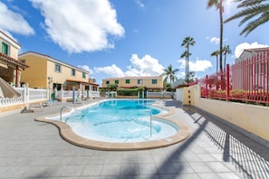 Una piscina con hidromasaje, rodeada por una valla blanca, palmeras y edificios con techos rojos.