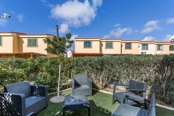 Un patio con mobiliario exterior, setos y una hilera de edificios con tejados de tejas rojas.