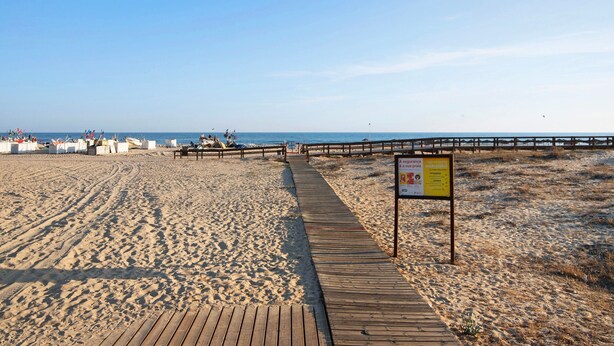 Una playa arenosa con un paseo marítimo de madera que conduce al agua, un puesto de salvavidas y un cartel.