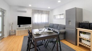 Un espacio habitable moderno con comedor, televisor de pantalla plana, sofá gris, refrigerador empotrado y área de cocina.
