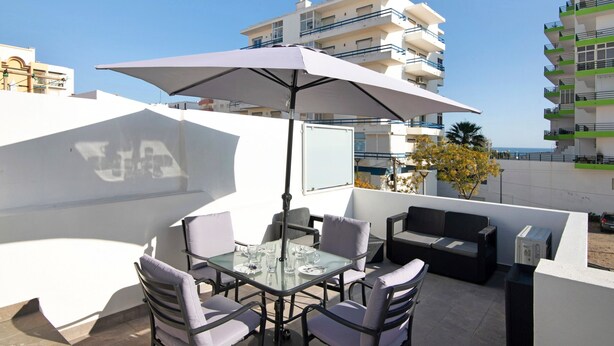 An outdoor patio with a table and chairs, a large umbrella, and a view of the sea and buildings. - Apartment "Telmus Villa 1 Andar" with Sea View, Private Terrace & Wi-Fi (Monte Gordo)