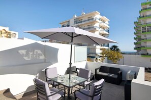 An outdoor patio with a table and chairs, a large umbrella, and a view of the sea and buildings.