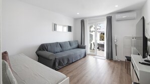A modern room with a grey sofa, a white bed, a desk with a computer, and a TV. - Apartment "Telmus Villa 1 Andar" with Sea View, Private Terrace & Wi-Fi (Monte Gordo)