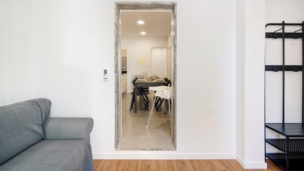 Een kamer met een grijze bank, een eettafel met stoelen en een metalen schapunit in het zwart.