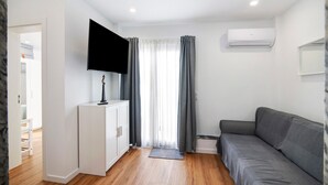 Een moderne woonkamer met een flatscreen televisie, een grijze bank, houten vloeren en een wit kastje.