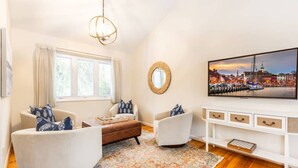 Un salon avec deux fauteuils blancs, un pouf marron, un tapis à motifs, un meuble TV blanc et un tableau représentant un paysage urbain.