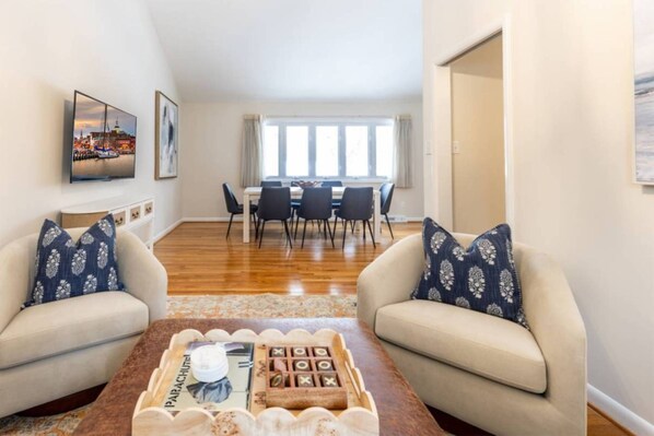 A living room with two sofas, a coffee table with a board game, a dining area with a table and chairs, and a large window with curtains.