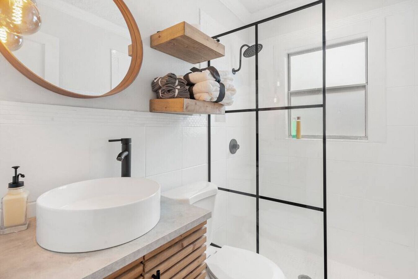 A modern bathroom with a round sink, a wooden shelf with toiletries, a black showerhead, and a large mirror.