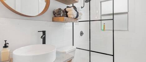 A modern bathroom with a round sink, a wooden shelf with toiletries, a black showerhead, and a large mirror.