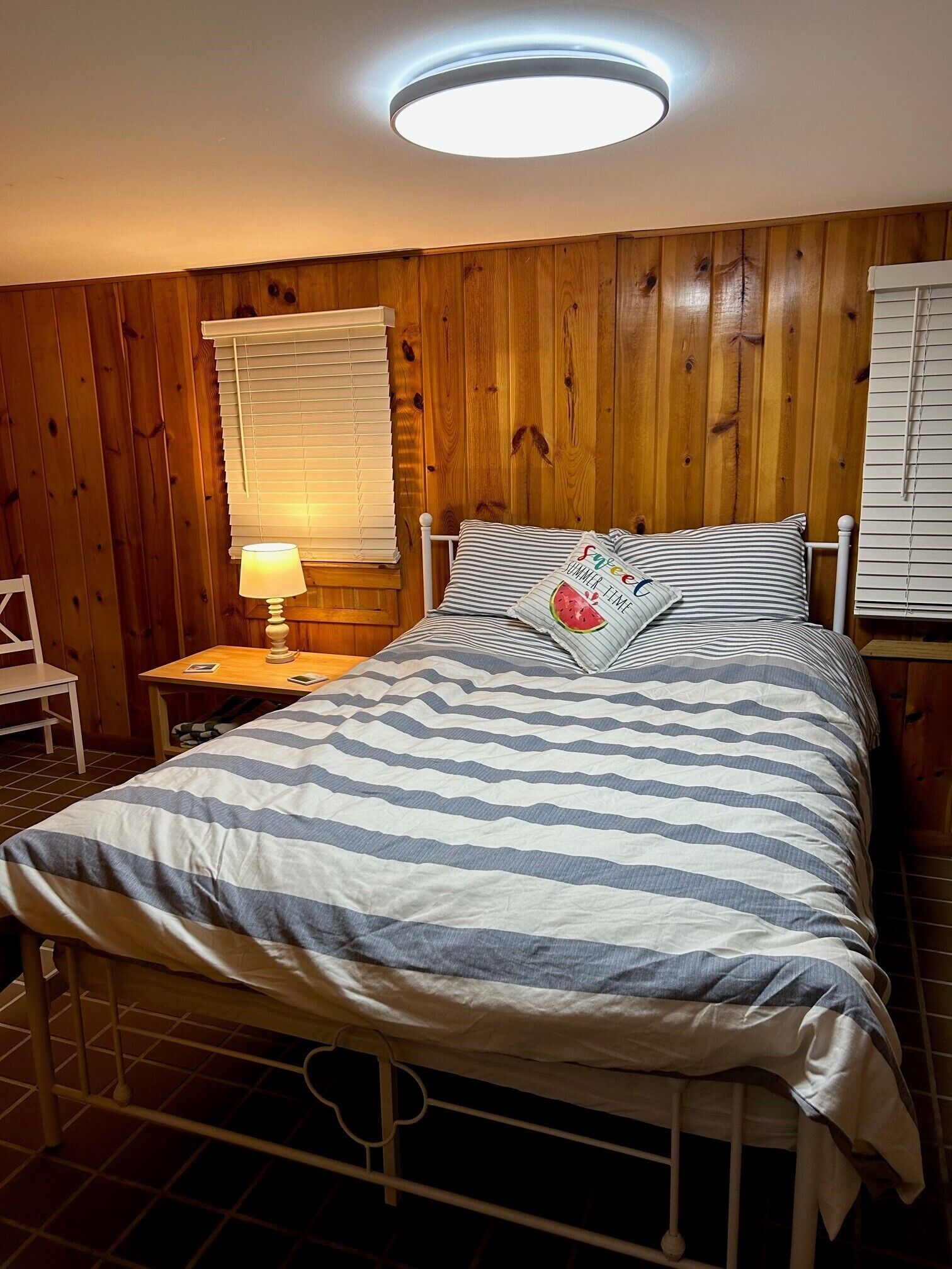 Een slaapkamer met een bed, houten wanden, een nachtkastje met een lamp en een raam met zonwering.