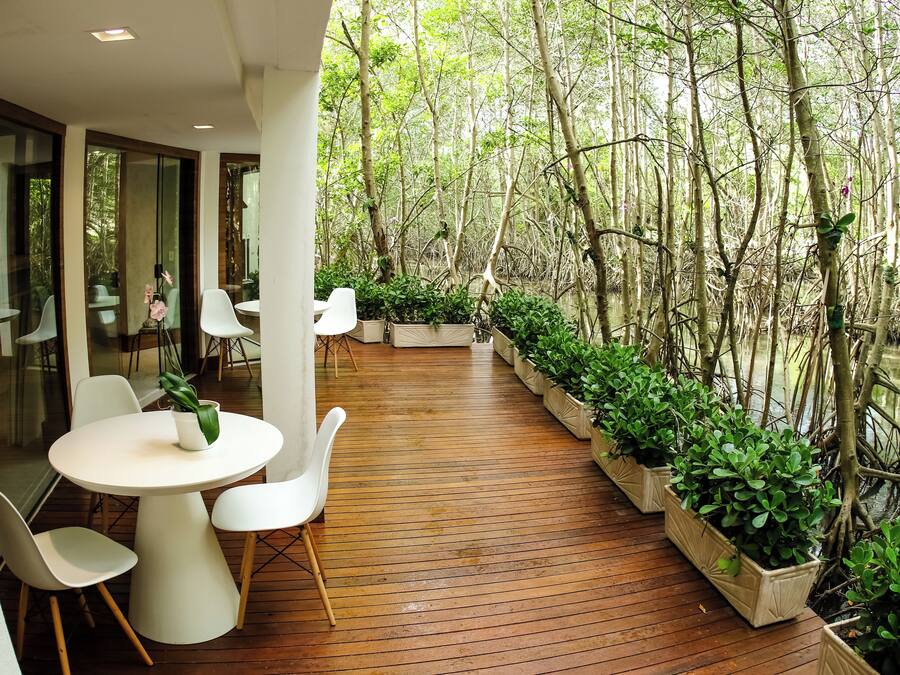 A wooden deck with white chairs and tables, surrounded by lush greenery and a water body.