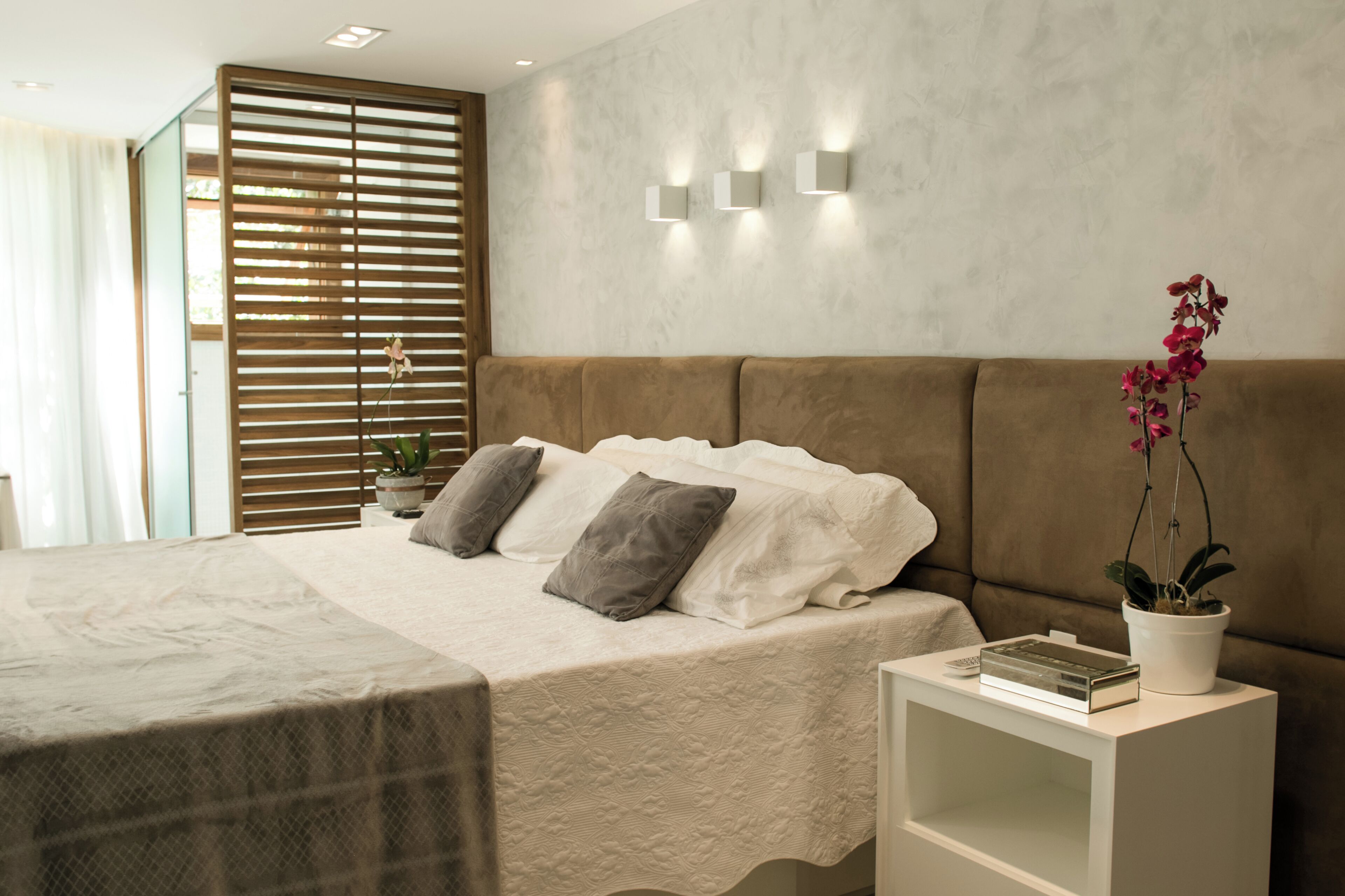 Quarto moderno com uma cama grande, mesa de cabeceira com um livro e um controle remoto, planta em vaso e janela com persianas.