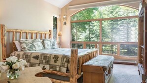 Un dormitorio con un ventanal grande, una cama de madera, un banco y un jarrón de flores.